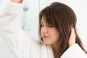 A woman in a white robe is closing her eyes in a bathroom while holding her hair with her right hand.