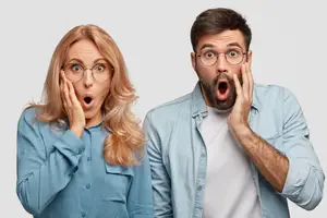 A man and a woman wearing glasses, posing for a picture with their mouths open and hands on their cheeks, standing against a white background.
