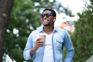 A smiling man is standing on the sidewalk, wearing a denim jacket, white shirt, and black sunglasses, holding a cup of coffee. Behind him, there are several trees, and the background is blurry, with a building in the distance. The man is also wearing earphones and has a beard and mustache.