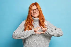 A woman with red hair is making a heart shape with her hands in front of a blue background.