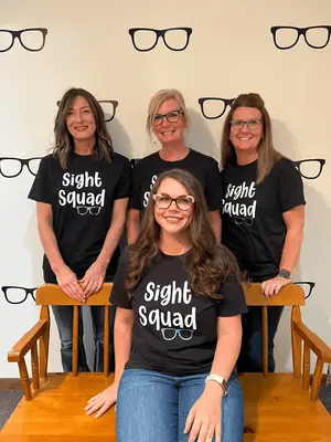 Five women wearing black Sight Squad shirts stand in front of a wall with eyeglasses printed on it. The woman in the middle is sitting on a wooden bench and smiling.