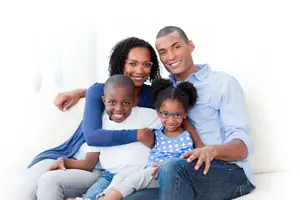 A smiling family of four, including a woman wearing glasses, a man in a light blue shirt, a boy in a white t-shirt, and a girl in a blue polka-dot dress, sitting together on a white couch.