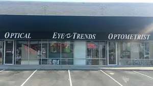 A storefront for Eye & Trends Optometrist, featuring a black awning with white lettering, a large glass window displaying various items, and a parking lot in front.