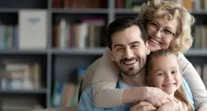 A man and a woman with a girl are sitting on a couch and smiling, and the woman is hugging the man and the girl.