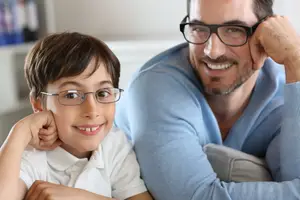 A man and a boy with glasses are smiling and looking at the camera. They are sitting on a couch in a room with a white wall, a shelf, and a file cabinet.