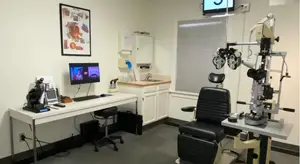A room with an eye examination chair and a desk with a computer monitor, a stool, a cabinet, and a monitor on the wall.