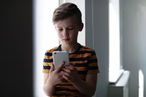 A young boy wearing a striped shirt is looking down at his phone in a room with windows and a white wall.