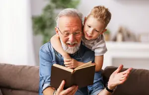 A grandfather reads a book to a boy on his lap, smiling and gesturing