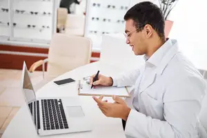 A man in a white lab coat holds a clipboard and pen while smiling at a laptop on a desk
