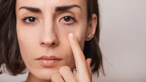 A woman with brown hair is touching her eye with her index finger while looking at the camera.