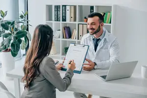 An adult woman is holding a clipboard and pen and speaking to a man in a white lab coat.