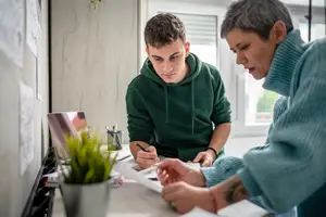 A boy and an older woman sitting at a desk, the boy appears to be writing something while the older woman looks at him.