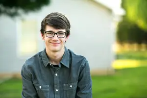 A smiling boy with glasses stands outside in front of a house with a tree in the background.