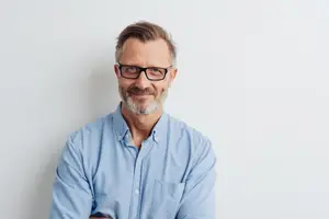 An older man with glasses and a beard is smiling and looking to the right while his arms are crossed.