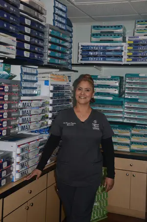 A woman in a uniform stands in a pharmacy, smiling and surrounded by neatly arranged medical supplies.