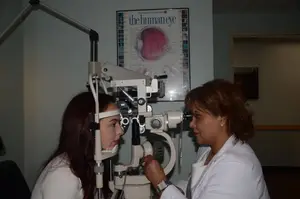 A woman looks into an ophthalmoscope while a female doctor adjusts it.