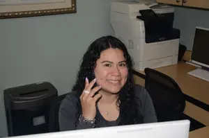 A woman with curly hair is sitting on a chair in front of a desk while holding a cell phone with a smile on her face.