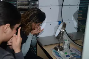 A woman is sitting in front of a mirror and looking at something while covering her mouth with her hands. Beside her is a man also sitting and looking at something. On the desk, there is a lamp, a mirror, and a box. Behind them are white storage boxes.