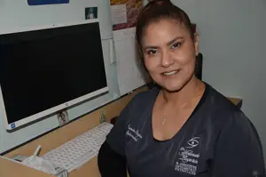 A woman smiling at the camera while standing in front of a desk with a computer and calendar on the wall