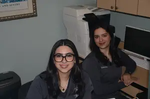 Two women are sitting in a room with a printer and a monitor on a desk in front of them.