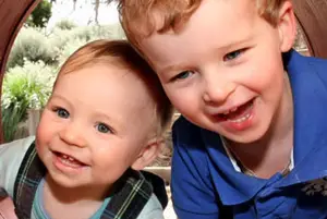Two young boys smiling and posing for a photo, one wearing a blue shirt and the other wearing a plaid shirt.
