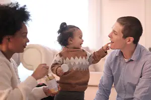 A woman, a man, and a toddler are sitting on the floor and the toddler is eating something from the jar the woman is holding.