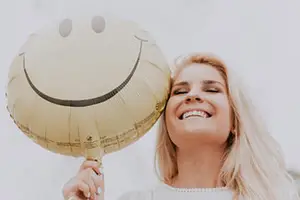 A woman with blonde hair is smiling while holding a smiley face balloon with her right hand