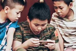 Three young boys are sitting on a couch and looking at their phones. They are all wearing casual clothing, and the boy in the middle has a camouflage shirt with a flag on it. The boy on the left is wearing a blue shirt with a pink design, while the boy on the right is wearing a white shirt with stripes. They are probably looking at a video or a photo on their phones.