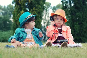 Two young children sitting on grass wearing hats, one holding a phone
