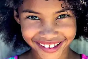 A young girl with curly hair and a bright smile is captured in a close-up shot, showcasing her vibrant green eyes and dimples on her cheeks. She is wearing a pink top with a white collar and has a mole on her left cheek. Her hair is messy and curly, and she is likely in a studio or a room with a light source behind her.