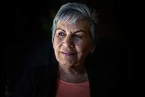 An elderly woman with gray hair and wrinkles on her face is smiling while looking forward in a dark room