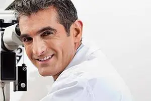 A smiling male doctor with a white coat looking through an ophthalmoscope