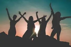 Five women are standing on a mountain and raising their hands in the air as the sun sets in the background.