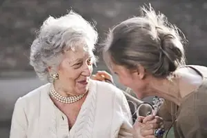 An elderly woman with white hair wearing a pearl necklace is smiling while a woman holding a stethoscope is talking to her.