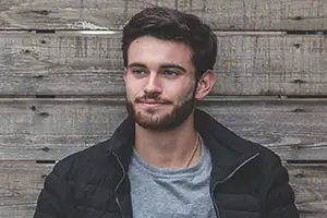 man with beard wearing black jacket and gray shirt standing against wooden wall