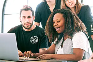 A group of people sitting in front of a laptop. A woman in the front is smiling and looking at the laptop. The man on her left is looking at the laptop while the other two people are looking at something else. There is a glass window behind them.