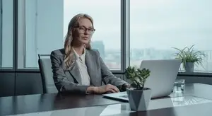 A woman in glasses is sitting at a desk in an office, working on her laptop, with a plant and a glass of water nearby, and a cityscape visible through the window.