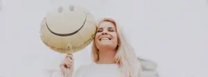 A woman with blonde hair is smiling and holding a smiley face balloon