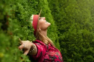 A woman wearing a hat and a pink floral shirt is leaning against a green bush with her head tilted upwards.
