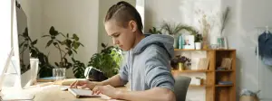 A teenager wearing a hoodie is sitting at a desk typing on a computer keyboard in a room with a wooden shelf and plants