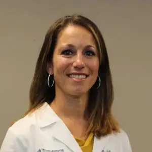 A smiling woman wearing a white lab coat and yellow shirt, standing in front of a light-colored wall.