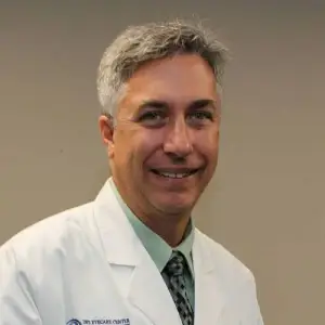 Dr. John DeSimone, an eye doctor with gray hair and a white coat, is smiling and posing for a photo.