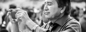 A black and white photo of a man taking a picture with a cellphone while standing in a crowd of people