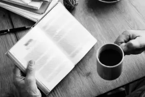 Image of two hands holding a book and a cup of coffee on a wooden table.