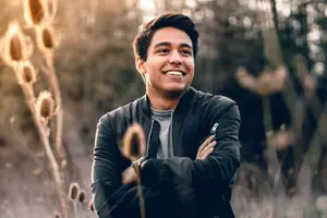 A smiling man in a black jacket standing in a field with his arms crossed