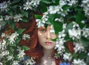 A woman with red hair is surrounded by white flowers, looking straight ahead.