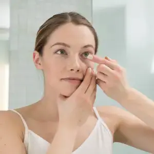 A woman applies eye cream to her eye in a bathroom mirror.