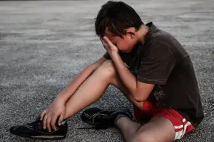 A young boy is sitting on the ground with his head on his hand and his eyes closed