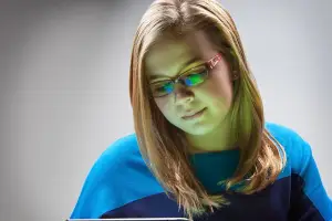 A young girl with long blonde hair wearing glasses and a blue sweater is looking down at a tablet