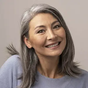 A smiling woman with gray hair and brown eyes wearing a blue shirt poses for a photo against a light gray background